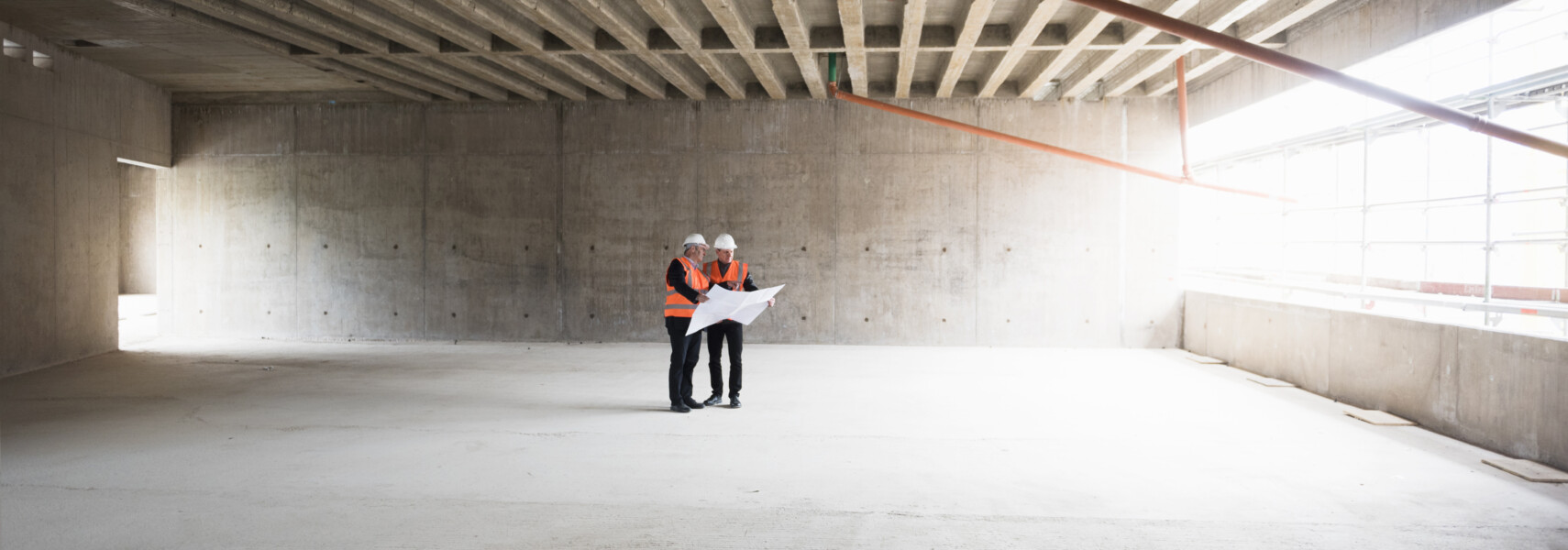 Two Men With Plan Wearing Safety Vests Talking In Building Under Construction
